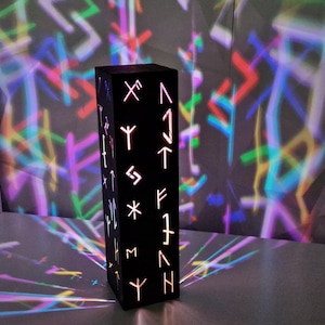 May include: A black wooden lamp with a geometric design and runes carved into the surface. The lamp casts a colorful light pattern on the wall behind it.