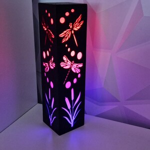 May include: A black wooden lamp with a geometric design and a dragonfly motif. The lamp is illuminated with pink and blue lights.