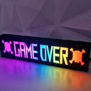 May include: A black light-up sign with the words "GAME OVER" in white and two skull and crossbones on either side. The sign is illuminated with rainbow colors.