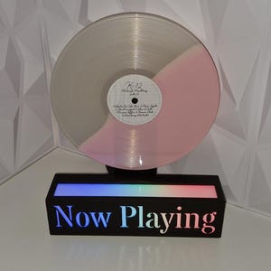 May include: A vinyl record with a pink and clear design is displayed on a black stand with the words "Now Playing" in a gradient colour scheme. The record is partially pink and has a white label. The stand has a colourful light display.