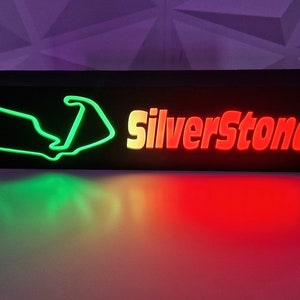 May include: A black wooden sign with a green outline of the Silverstone racetrack and the word "Silverstone" in red letters.