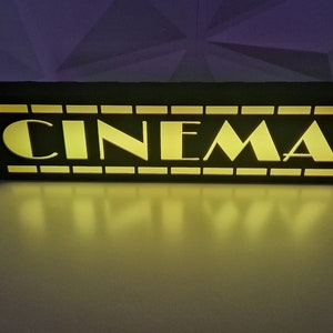 May include: A black wooden sign with the word "CINEMA" in yellow letters. The sign is lit up from behind, creating a warm glow.