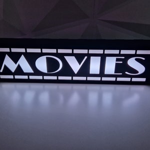 May include: A black rectangular light box with the word "MOVIES" in white, illuminated letters. The light box has a film strip design around the edges. The light is bright and casts a glow on the surface below.