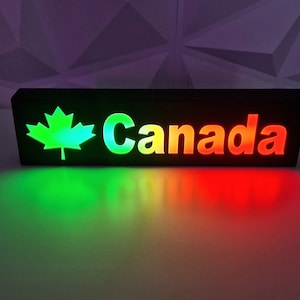 May include: A black wooden sign with the word "Canada" in white letters and a green maple leaf. The sign is illuminated with red, orange, yellow, and green lights.