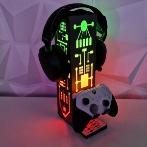 May include: A black gaming headset stand with a built-in controller holder. The stand has a green and red LED light design.
