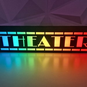 May include: A black wooden sign with the word "THEATER" in white letters. The sign is illuminated with a rainbow of colors, from blue to red.