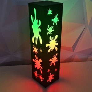 May include: A black wooden lamp with a rectangular shape and a design of green and red paint splatters cut out of the wood. The lamp is lit from the inside, casting a green and red glow.