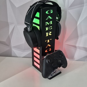 May include: Black gaming headset stand with a green and red light display. The stand has the word "GAMER TAG" printed on it. A black video game controller is resting on the stand.