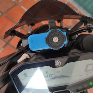 Puede incluir: Un soporte de teléfono para motocicleta azul con un botón pulsador y una cerradura. El soporte está fijado al manillar de una motocicleta.