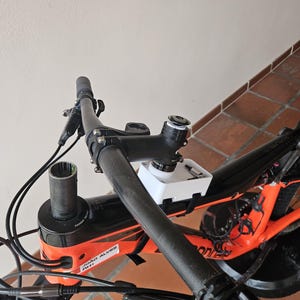 May include: A mountain bike cockpit featuring a black handlebar, a stem, and a white 3D-printed storage box mounted on the frame. The frame is orange and black with a sticker reading JOAO ALVES. Brake levers, cables, and a headset are visible.