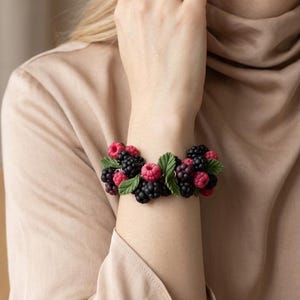 May include: A bracelet featuring a cluster of realistic miniature blackberries and raspberries interspersed with small green leaves. The fruit elements are dark purple, black, and bright pink, arranged in a dense, continuous band around the wrist.