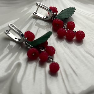 May include: Silver-tone earrings featuring clusters of red berry-like beads and green leaf accents. The earrings have a hinged closure and are displayed on a white, textured surface. The design evokes a natural, fruity theme.
