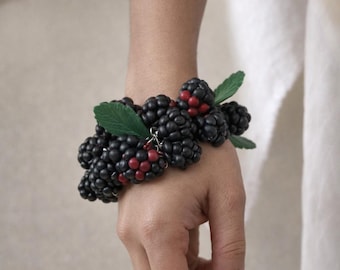 May include: A bracelet featuring clusters of black and red berry beads with green leaf accents. The bracelet is worn on a wrist, showcasing the handmade jewelry's naturalistic design. The berries are approximately 1 inch in diameter.