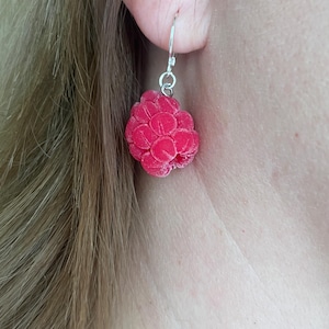Op de afbeelding: Een hangende oorbel met een realistische, getextureerde framboosbedel in een levendige roze-rode kleur. De fruitvormige bedel hangt aan een eenvoudige zilverkleurige metalen haak, wat zorgt voor een gedetailleerd en lichtgewicht accessoire.