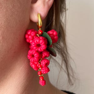 以下が含まれることがあります： 鮮やかなピンク色のラズベリーのイヤリング。イヤリングは、詳細なラズベリーの形がクラスター状になっており、緑の葉と金色のアクセントが付いています。イヤリングは金色のフープに取り付けられています。