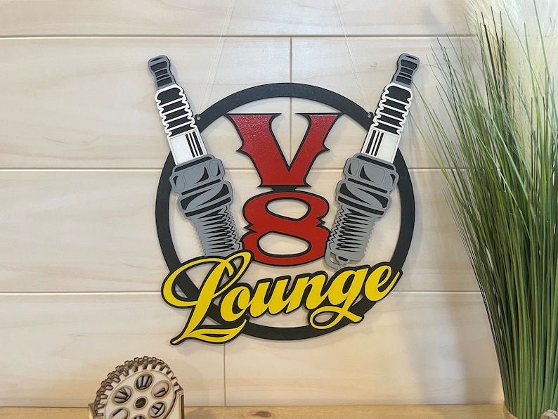 May include: A black metal circle with a red and yellow "V8 Lounge" sign. Two gray spark plugs are crossed behind the sign.