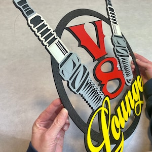 May include: A black and yellow sign with the text "V8 Lounge" in a yellow script font. The sign is decorated with two spark plugs, one on each side of the text. The spark plugs are gray and black with white accents.