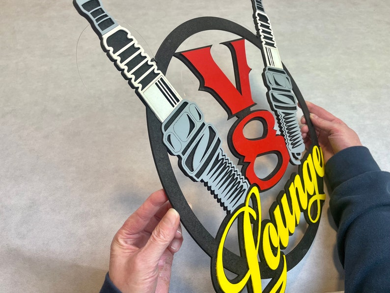 May include: A black and yellow sign with the text "V8 Lounge" in a yellow script font. The sign is decorated with two spark plugs, one on each side of the text. The spark plugs are gray and black with white accents.