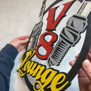 May include: A black, red, and yellow wooden sign with the text "V8 Lounge" and a stylized image of three spark plugs.