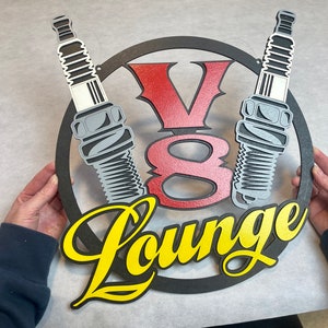 May include: A black metal sign with a red and white V8 logo and two spark plugs. The sign says "Lounge" in yellow letters.