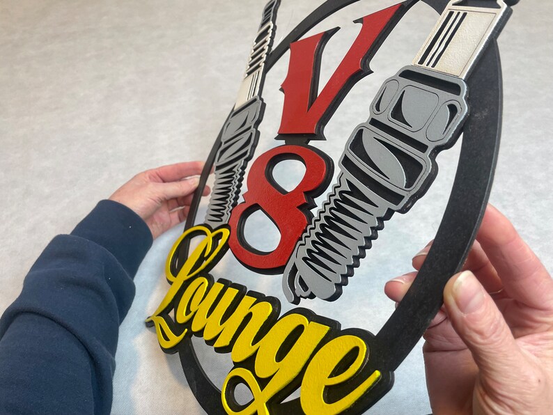 May include: A black, red, and yellow wooden sign with the text "V8 Lounge" and a stylized image of three spark plugs.