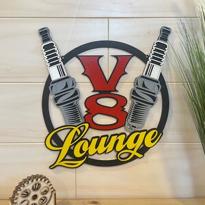 May include: A black metal circle with a red and yellow "V8 Lounge" sign. Two gray spark plugs are crossed behind the sign.
