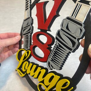 May include: A black, round sign with a red "V8" and yellow "Lounge" text. Two silver spark plugs are attached to the sign.