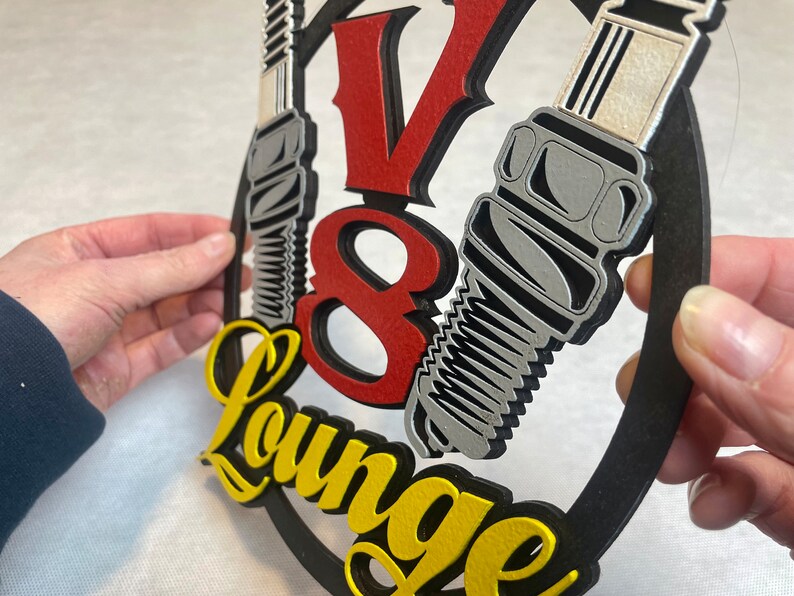 May include: A black, round sign with a red "V8" and yellow "Lounge" text. Two silver spark plugs are attached to the sign.