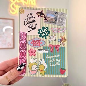 May include: A decorative Kindle case featuring various stickers and text. The stickers include a whale, a camera, a four-leaf clover, a unicorn, and the words "The Couch Club," "Slow Reader Club," and "Kindle Girly." The case also has the text "happiest: with my kindle."