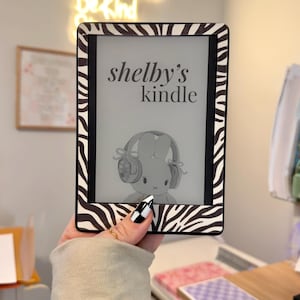 May include: An e-reader device featuring a black and white zebra print frame. The screen displays the text "shelby's kindle" above a graphic illustration of a cartoon rabbit wearing headphones. The device has a black border around the display.