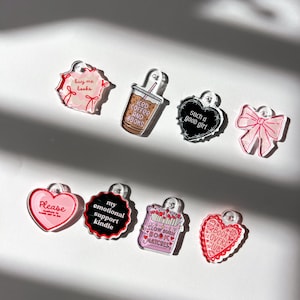 May include: A collection of acrylic charms in various shapes, including hearts, bows, and a coffee cup. Each charm features a unique design with text such as "buy me books," "iced coffee and books," and "book lovers club." The charms are in shades of pink, red, black, and brown.