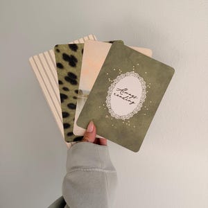 May include: A set of four rectangular cards with rounded corners. The front card is olive green with a white lace-style oval frame containing the text Always reading. Other cards feature a leopard print, vertical stripes, and a landscape scene.