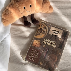 May include: A plush croissant toy with a smiling face and brown legs, positioned next to a clear tablet case featuring a collage of a coffee drink, sunglasses, a brown bunny, and text that reads READ MORE BOOKS, BOOK LOVER, and SLOW DOWN YOU'RE DOING FINE.