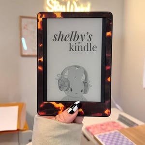 May include: A Kindle e-reader with a tortoiseshell-patterned case. The screen displays "shelby's kindle" and a cartoon bunny wearing headphones. The e-reader is held in a hand with a black and white checkered nail design.