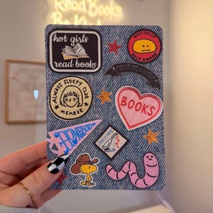May include: A rectangular denim-textured card featuring various embroidered-style patches including a book with the text hot girls read books, a yellow face with the text BRUTALLY SELF AWARE, a heart with BOOKS, a pink pennant, and a cartoon worm.
