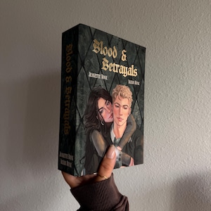 May include: A hardcover book titled Blood & Betrayals by Jeanette Rose and Alexis Rune. The cover features a dark green diamond-patterned background with an illustration of a dark-haired person embracing a blonde person in a black jacket.