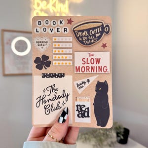 May include: A collection of decorative stickers on a brown background. The stickers include phrases like "Book Lover," "Drink Coffee & Do All The Things," and "The Slow Morning." Other stickers feature a black cat, a four-leaf clover, and a coffee cup.
