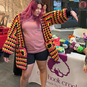 May include: A colorful, handmade crochet cardigan in black, yellow, orange, and pink. The cardigan has a geometric pattern and pockets. The image also shows a table with various crochet items, including stuffed animals, and the sign "hook crochet".