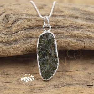 May include: A pendant necklace featuring an elongated, dark olive green, textured stone encased in a smooth silver bezel setting. The pendant hangs from a delicate silver chain, showcasing a raw, organic appearance with a glossy finish.
