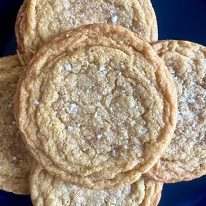 May include: A close-up shot of several golden-brown cookies, sprinkled with coarse white salt crystals. The cookies are round with slightly textured surfaces and are stacked on top of each other. The background is a dark blue.