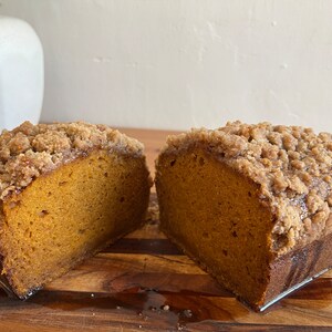 K&ouml;nnte beinhalten: Nahaufnahme eines K&uuml;rbis-Laibkuchens mit Streusel, halbiert und auf einem Holzbrett pr&auml;sentiert.