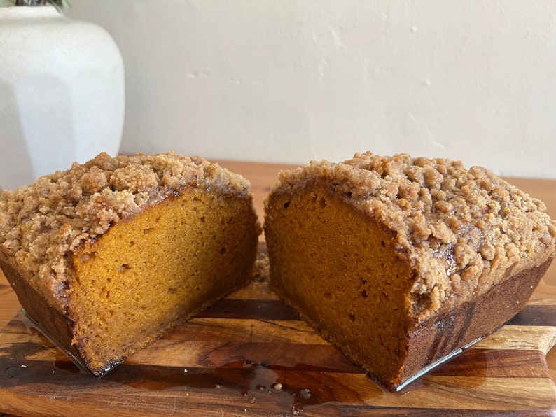 K&ouml;nnte beinhalten: Nahaufnahme eines K&uuml;rbis-Laibkuchens mit Streusel, halbiert und auf einem Holzbrett pr&auml;sentiert.