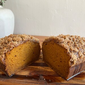 K&ouml;nnte beinhalten: Ein Laib K&uuml;rbisbrot mit Streusel-Topping, halbiert und auf einem Holzbrett pr&auml;sentiert.