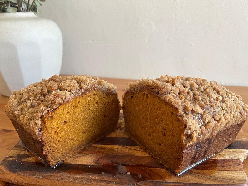 K&ouml;nnte beinhalten: Ein Laib K&uuml;rbisbrot mit Streusel-Topping, halbiert und auf einem Holzbrett pr&auml;sentiert.