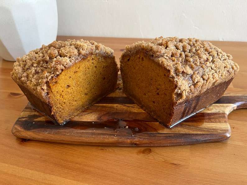 K&ouml;nnte beinhalten: Ein Laib K&uuml;rbisbrot mit Streusel-Topping, halbiert und auf einem Holzbrett pr&auml;sentiert.