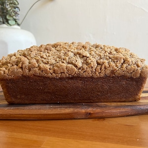 May include: A loaf of bread with a crumb topping. The bread is on a wooden cutting board.