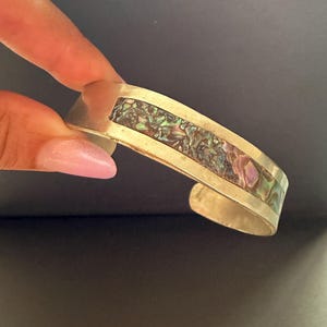 Op de afbeelding: Een zilverkleurige armband met een centrale horizontale inleg van iriserende abalone-schelp. De schelp vertoont een mozaïek van glinsterende groen-, paars- en blauwtinten, gevat in een gladde, gepolijste metalen band met een open achterkant.