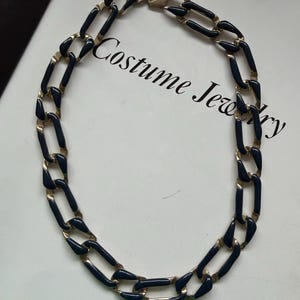 Op de afbeelding: Een goudkleurige ketting met langwerpige ovale schakels en donkerblauwe emaille inleg. De ketting heeft een veilige sluiting en wordt getoond op een witte ondergrond met de tekst Costume Jewelry in zwarte letters afgedrukt.