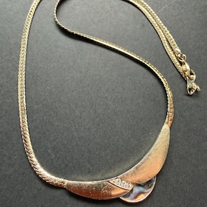 Op de afbeelding: Goudkleurige ketting met een gebogen hanger. De hanger heeft een cirkelvormige iriserende inleg en is versierd met kleine, heldere kristallen. De ketting heeft een textuur, geweven ontwerp en een sluiting. De ketting wordt op een donkere ondergrond getoond.
