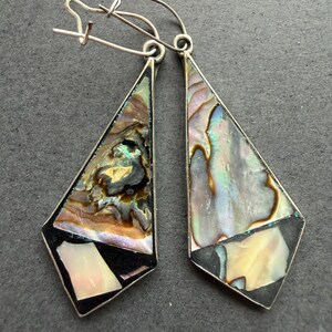 Op de afbeelding: Een paar hangende oorbellen met langwerpige driehoekige monturen van zilverkleurig metaal. Elke oorbel is ingelegd met iriserende abalone schelpsegmenten in tinten groen, blauw en roze, geaccentueerd door effen zwarte secties en kleine witte schelpstukjes.